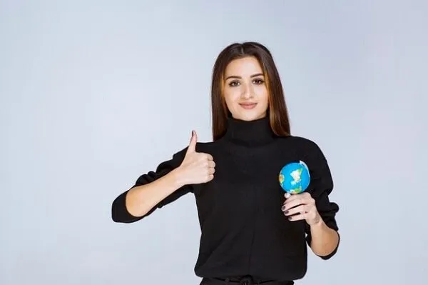  Молодая девушка держит мини глобус и показывает пальцем знак класс. 