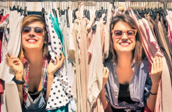 Две молодых женщины в солнцезащитных очках выглядывают из-за вешалок с одеждой