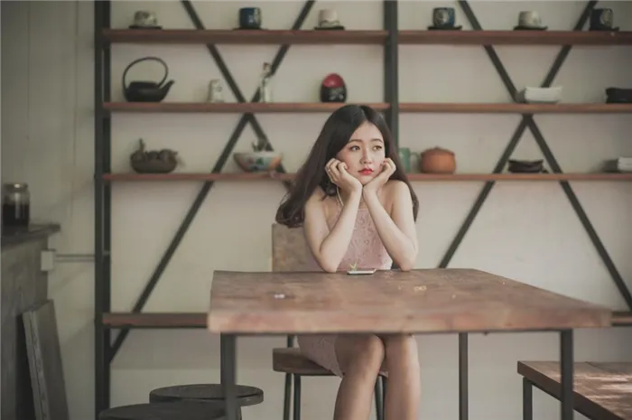 Девушка сидит за столом со скучающим видом на фоне полок с кухонной утварью